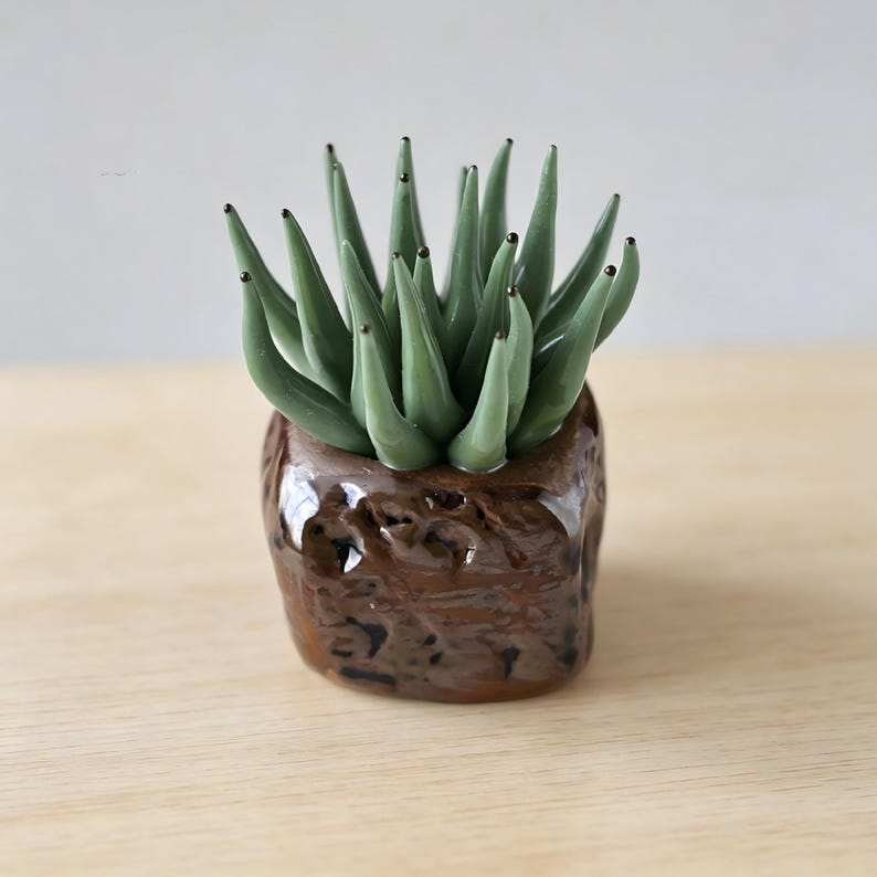 May include: A small glass sculpture of a succulent plant with pointed, sage green leaves featuring dark tips. The plant is set into a textured, dark brown, square-shaped base with a glossy finish that mimics the appearance of natural soil.