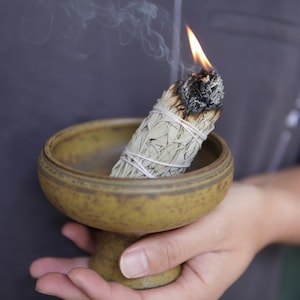 May include: A burning white sage bundle, tied with white string, emits smoke. The sage is held in a small, round, pedestal-style ceramic bowl with a yellow-brown glaze. The flame is bright orange against the sage's dark, charred end.