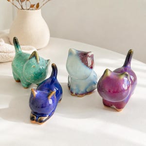以下が含まれることがあります： 様々な色の4つの小さな陶器製の猫の置物。1つは緑色の斑点模様、1つは濃い青色、1つは濃い紫色の斑点がある水色、もう1つは鮮やかな紫色です。それぞれの猫には尖った尻尾と耳があり、滑らかで光沢のある仕上がりになっています。