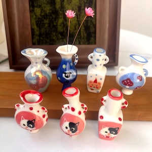 Könnte beinhalten: Eine Sammlung von Miniatur-Keramikvasen in verschiedenen Designs. Die Vasen weisen handgemalte Details auf, darunter Katzenmotive, Tupfen und Cartoonfiguren. Einige Vasen enthalten zarte rosa Blüten, die der Präsentation einen Hauch von Farbe und Charme verleihen.