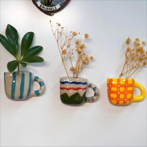 May include: Three ceramic teacup-shaped planters with handles are mounted on a white surface. One is striped blue and white, another has red, white, and green stripes, and the third is yellow with an orange grid pattern. Each contains plants.