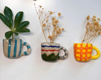 Op de afbeelding: Drie keramische theekopvormige plantenbakken, elk met een handvat, zijn op een wit oppervlak gemonteerd. Eén is blauw en wit gestreept, een andere heeft rood, wit en groene strepen, en de derde is geel met een oranje ruitpatroon. Elke bak bevat planten.