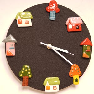 Puede incluir: Un reloj de pared caprichoso con una esfera negra, con casas y setas de cerámica coloridas como marcadores de hora. Las manecillas del reloj son blancas. El diseño es lúdico y decorativo.