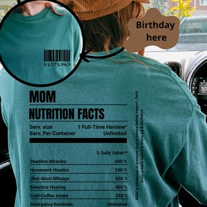 以下が含まれることがあります： ターコイズブルーのTシャツに、「MOM」と書いてある栄養成分表示ラベルが付いています。ラベルには、サービングサイズとして「フルタイムヒロイン」と記載されています。ラベルには、「デッドラインミラクル」や「ウーバーマママイレージ」など、ユーモラスな「栄養素」のリストが記載されています。