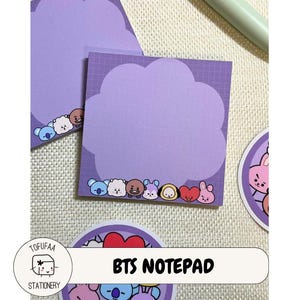 May include: Purple BTS notepad with a cloud-shaped border and grid pattern. The bottom border features cartoon characters. The notepad is accompanied by a round sticker with a cartoon character and the text "BTS NOTEPAD".