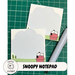 May include: Two Snoopy notepads with a cloud-shaped design and a Snoopy illustration in his doghouse. The notepads have a light gray background and a green border. A pen rests on the green grid background.