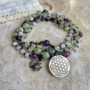 May include: A beaded necklace featuring green, purple, and white gemstones. The necklace has a silver lotus charm and a silver pendant with a geometric design. The beads are strung on a dark cord.
