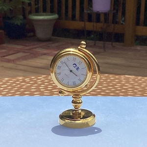 May include: A gold-toned globe-shaped desk clock with a white face, black Roman numerals, and a blue logo. The clock sits on a light blue surface decorated with a blue butterfly and a colorful floral arrangement of pink, yellow, and purple flowers.