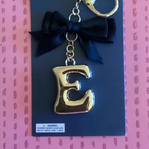 May include: A gold-colored letter "E" keychain with a black bow and a gold-colored clasp. The keychain is attached to a black card with the text "iRON ORCHID" and a warning about small parts.
