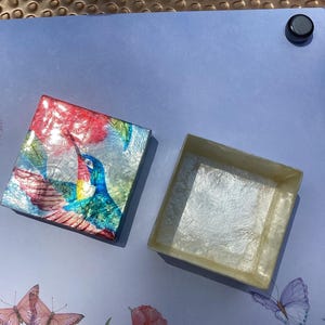 May include: A square gift box with a separate lid featuring a vibrant, glossy illustration of a hummingbird in shades of blue, green, and red. The interior of the box and the underside of the lid have a textured, off-white, pearlescent finish.