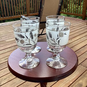 Op de afbeelding: Vier vintage glazen kelken met een tarwe- en bloemenontwerp in zwart en zilver. De kelken staan op een kleine, ronde, donkerbruine tafel met een gedraaide poot. De tafel staat op een houten terras.