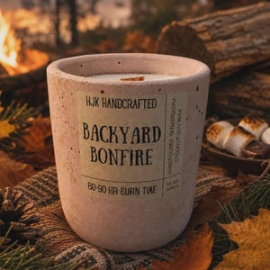 May include: A cylindrical concrete candle vessel with a light beige speckled finish. The label reads HJK HANDCRAFTED BACKYARD BONFIRE, 80-90 HR BURN TIME, HANDPOURED IN NEBRASKA, COCONUT SOY WAX, 12 OZ, 340 G. The candle features a wooden wick.