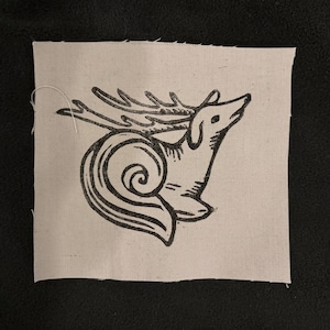 May include: A square, off-white fabric patch featuring a black illustration of a deer-snail hybrid. The creature has deer antlers and a snail shell, with a stylized design. The patch is on a black background.