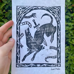 May include: Black and white linocut print depicting a dog with a sword through its back, fighting a snake. The print is framed by an ornate border with floral and geometric elements. The artist's signature is in the lower right corner.