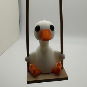 May include: A white goose figurine with orange beak and black eyes sits on a brown wooden swing set. The swing is suspended by a red and white string.