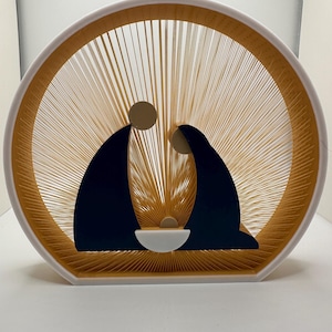 May include: A decorative nativity scene featuring a circular design with a white frame and gold-colored string radiating from the center. Silhouettes of Mary, Joseph, and the baby Jesus are in the center, creating a religious theme.