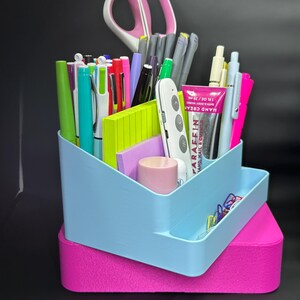 May include: A two-tiered desk organizer in light blue and pink, filled with pens, pencils, scissors, sticky notes, and a tube of hand cream. The organizer has multiple compartments for stationery and office supplies.