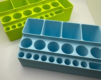 May include: Two plastic desk tidies in lime green and light blue. Each tidy has multiple compartments, including square and round holes of varying sizes, designed for storing pens, pencils, and other stationery. The tidies are on a white surface.