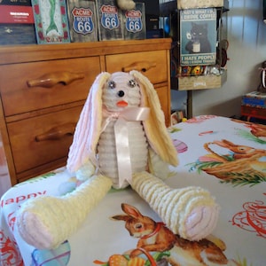 May include: A handmade plush bunny rabbit with long, floppy ears and a pink bow. The bunny is white with yellow and pink accents, and is sitting on a floral tablecloth. The bunny's eyes are embellished with rhinestones.