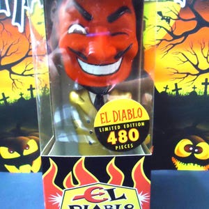 May include: A collectible bobblehead figure of a red-faced devil with a black goatee, wearing a gold suit and black tie. The figure is encased in a clear plastic box with a yellow sticker reading EL DIABLO LIMITED EDITION 480 PIECES.