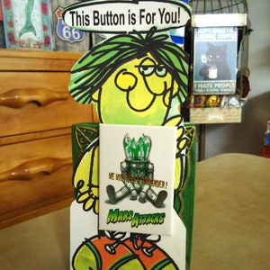 May include: A die-cut cardboard display featuring a cartoon character with green hair and a speech bubble that reads This Button is For You! A rectangular pinback button is attached to the front, depicting a Mars Attacks alien with the text WE WILL NEVER SURRENDER! and MARS ATTACKS.