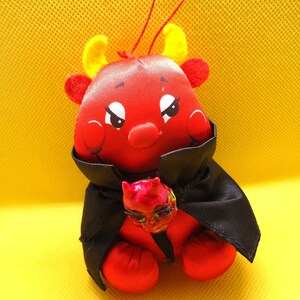 May include: A plush red devil toy with yellow horns, wearing a black satin cape. A small, detailed red devil face charm is attached to the front of the cape. The toy features a red string loop at the top for hanging and has a smooth, shiny fabric finish.