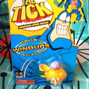 May include: A packaged "The Tick" action figure. The packaging is yellow and blue, featuring the character's face and the text "Wacky Windups Action". The toy is a yellow submarine with a purple top.