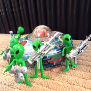 May include: A group of bright green alien figurines holding silver toy rifles, positioned in front of a round, blue and red metal flying saucer toy with a clear dome top and the text X-7 printed on the side of the metallic craft.