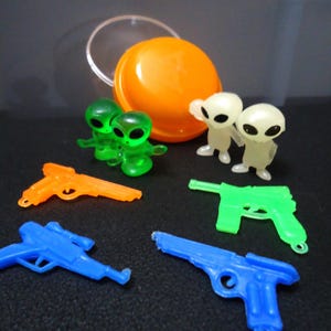 May include: A collection of plastic toys featuring alien figures and toy guns. The aliens are in green and glow-in-the-dark white. The toy guns are in blue, green, and orange. An orange container is also present.