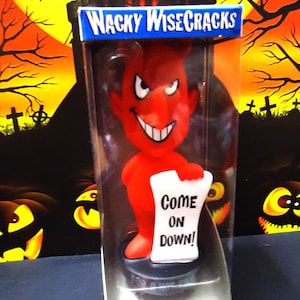 May include: A red devil figurine with a smiling face and white teeth, holding a scroll that reads "Come On Down!" The figurine is packaged in a clear plastic box with the words "Wacky WiseCracks" on top. The background features Halloween-themed imagery.