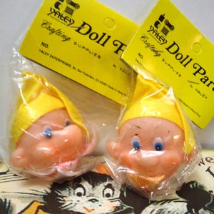 May include: Two vintage doll heads with yellow caps and peach-colored skin, packaged in clear plastic. The doll heads have blue eyes and rosy cheeks. Yellow tags with the text "Yaley Doll Parts Crafting Supplies" are visible in the background.