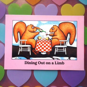 May include: A pink greeting card featuring a cartoon illustration of two squirrels dining outdoors. The squirrels are seated at a table with a red and white checkered tablecloth. The card reads "Dining Out on a Limb."