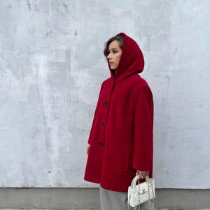 May include: A vibrant red hooded coat featuring black buttons and pockets. The coat is worn with a grey skirt, black shoes, and a white quilted handbag with a gold chain strap. The background is a textured grey wall.