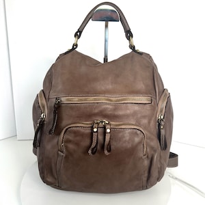 Könnte beinhalten: Ein brauner Lederrucksack mit mehreren Reißverschlussfächern und einem oberen Griff. Die Tasche hat ein abgenutztes Aussehen mit sichtbaren Nähten und messingfarbener Hardware. Das Design umfasst zwei Fronttaschen und zwei Seitentaschen.