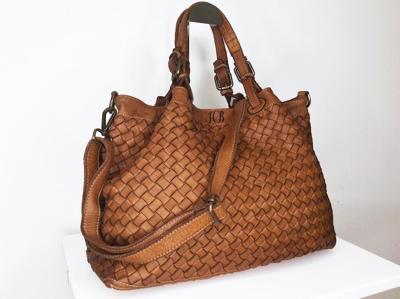 Pu&ograve; includere: Una borsa tote in pelle intrecciata marrone con un manico superiore e una lunga tracolla. La borsa ha un design intrecciato e le iniziali "JCB" sono impresse sul davanti.