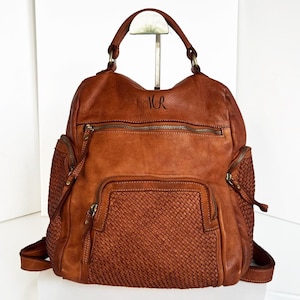 Peut inclure: Un sac à dos en cuir marron avec un détail tissé sur le devant. Le sac à dos a une poignée supérieure et des bretelles réglables. Les initiales "M.R." sont gravées sur le devant du sac.