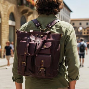 May include: A dark burgundy leather backpack featuring two front pockets with buckle closures, a top zipper, and adjustable shoulder straps. The bag has a structured rectangular shape with visible stitching and antique brass-toned metal hardware.