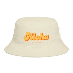 May include: A cream-coloured bucket hat featuring the word "Aloha" in orange, retro lettering. Beneath, the text reads "GOLDEN HOUR BY TAI". The hat is made of a soft material and has a wide brim, perfect for sun protection.