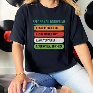 May include: Black t-shirt with the text "BEFORE YOU BOTHER ME" and a numbered checklist: "1. IS IT PLUGGED IN?", "2. IS IT TURNED ON?", "3. ARE YOU SURE?", and "4. SERIOISELY, GO CHECK". The shirt is worn by a person.