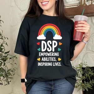 May include: Black t-shirt with a rainbow and the text "DSP Empowering Abilities, Inspiring Lives." The shirt has a colorful design with hearts. The person is wearing blue jeans and holding a red cup with a straw.
