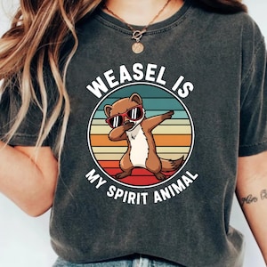 Puede incluir: Camiseta gris oscuro con un dibujo animado de una comadreja con gafas de sol y haciendo dab, sobre un fondo a rayas. El texto "WEASEL IS MY SPIRIT ANIMAL" se muestra en un círculo.