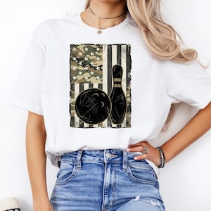 May include: A white short-sleeve t-shirt featuring a rectangular graphic on the chest. The graphic displays a stylized American flag pattern with camouflage and stripes, overlaid with a black bowling ball and a single black bowling pin.