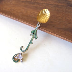 May include: A decorative spoon with a gold-colored shell-shaped bowl. The handle features a green and purple floral design with blue accents. The spoon is likely made of metal and is designed for decorative purposes.