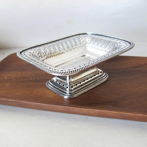 May include: A silver-colored, rectangular dish with a decorative, openwork border and a pedestal base. The dish sits on a wooden tray. The dish's rim features a repeating pattern, and the base has a series of small, evenly spaced holes.