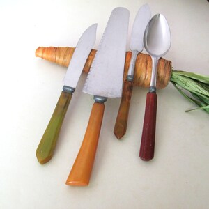May include: A set of four stainless steel utensils featuring distinct handles: a knife with a green handle, a serrated cake server with an orange handle, a spreader with a brown handle, and a spoon with a dark red handle, resting on a carrot.