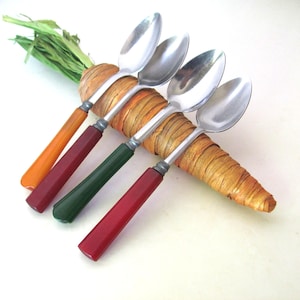 May include: Four stainless steel spoons with solid-colored handles in orange, maroon, dark green, and red. The spoons are arranged diagonally across a decorative, textured, carrot-shaped object made of wrapped natural fibers with green leafy tops.