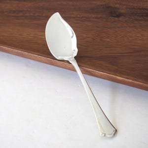 May include: A silver serving spoon with a leaf-shaped head and a long, slender handle. The spoon is resting on a dark wooden surface, with a white surface in the background. The spoon has a decorative edge.