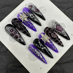 May include: A collection of artificial nails in various designs. Some nails are black, purple, and clear, with intricate silver and gold embellishments. The nails feature patterns and textures, including swirls and geometric shapes.