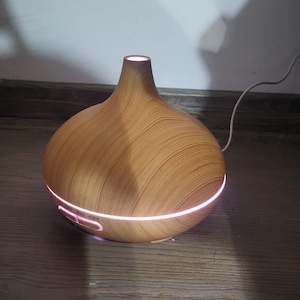 Könnte beinhalten: Ein ätherisches Öl Diffusor in Holzoptik mit rosa Licht an der Basis und Oberseite. Der Diffusor hat einen bauchigen Körper und einen schmalen Hals. Ein weißes Netzkabel ist angeschlossen.