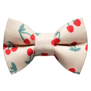 May include: A cream-colored bow tie with a red cherry print. The bow tie is made of fabric and has a classic bow shape.